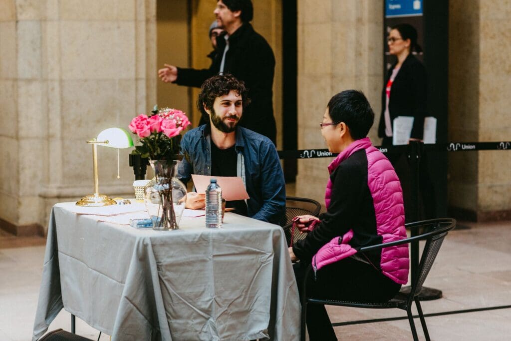 A bearded man faces a woman; they are sitting on the same side of a table that has a lamp and flowers.