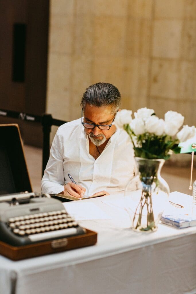 A man sits at a table with flowers and a typewriter, writing a poem on a sheet of paper
