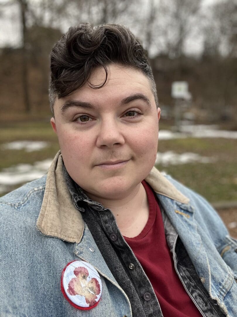 Photo of Tara Borin (they/them) standing outside in a denim jacket